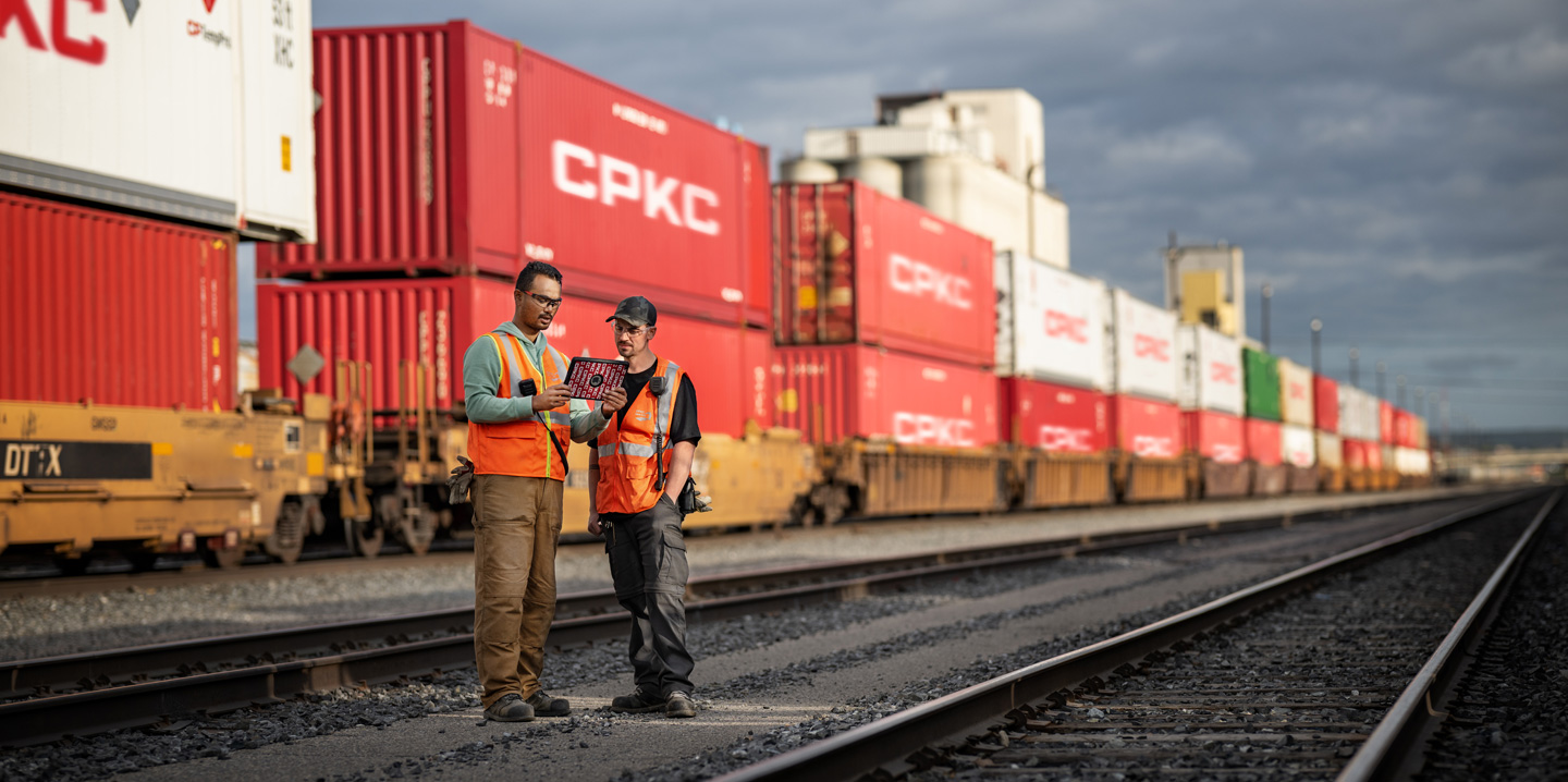 Two railway workers reference an iPad while standing in a train depot beside a stationary train carrying shipping containers